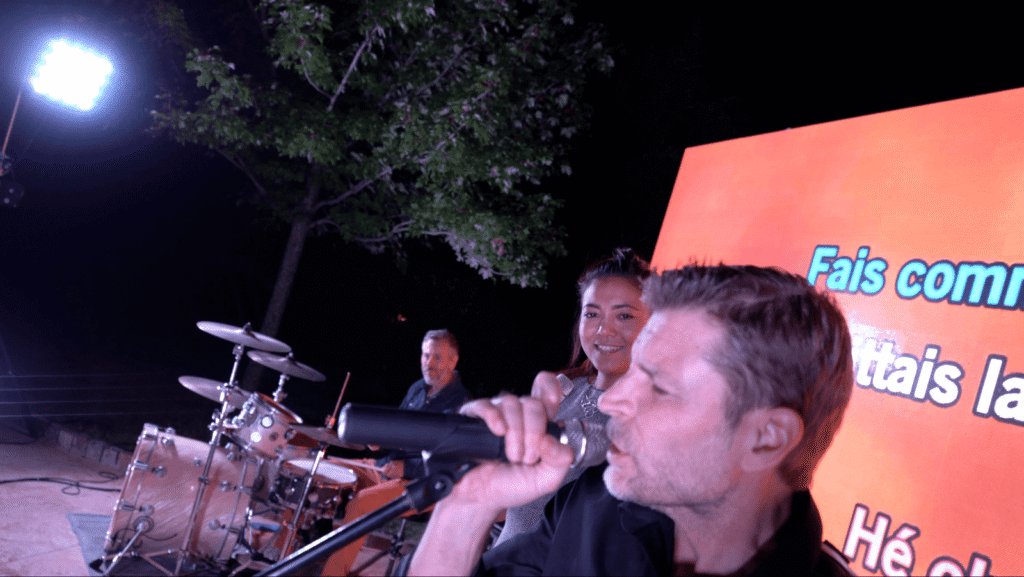 Un chanteur au micro devant la batterie, écran orange derrière