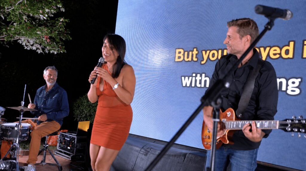 La chanteuse, le guitariste et le batteur sur scène devant les paroles projetées — image retouchée
