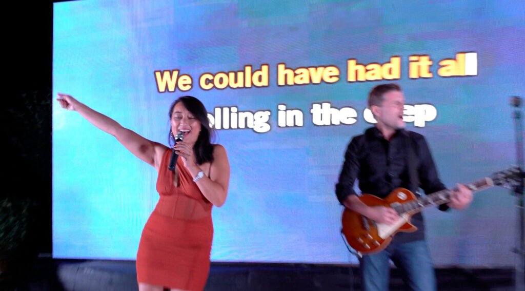 Une candidate en robe rouge chante bras tendu, guitariste derrière