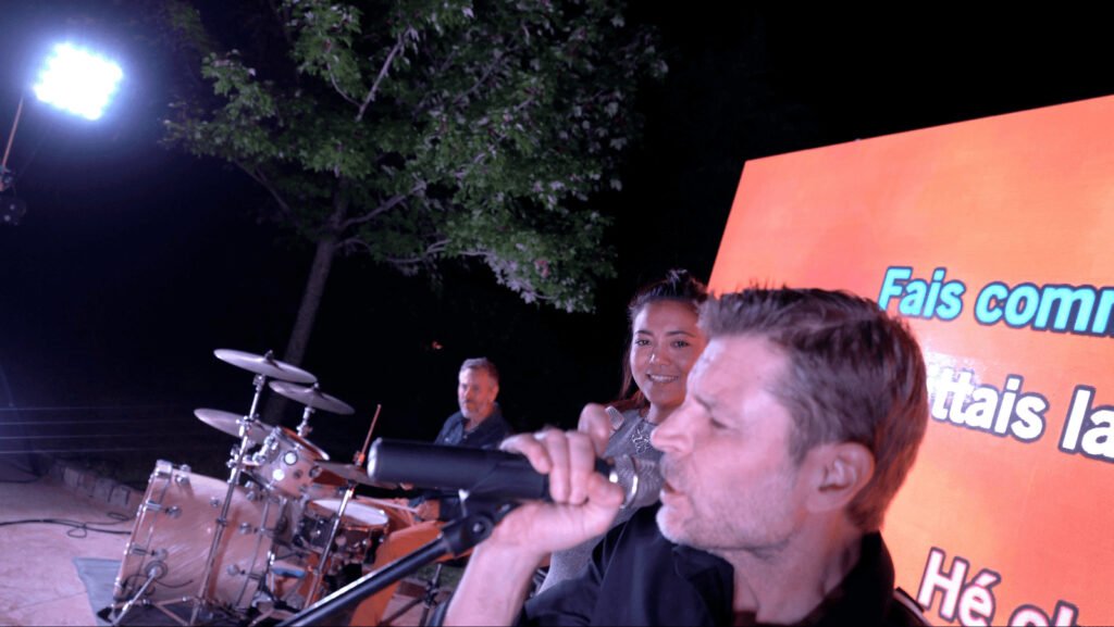 A singer at the mic in front of the drum kit, with an orange screen behind